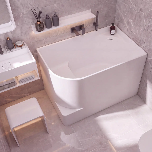 A modern freestanding bathtub in a minimalist bathroom setting with gray marble-look tiled walls and floor, a white vanity, and a small white stool.