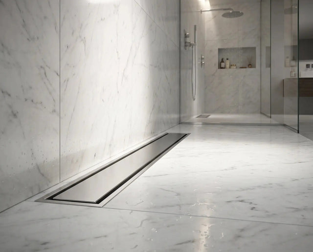 A brushed stainless steel linear channel floor drain with a removable cover plate, installed in a modern walk-in shower floor made of large white marble tiles, in a luxury bathroom.