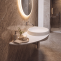 A modern wall-mounted white vessel sink in a bathroom with warm gray marble walls and floor, featuring an illuminated round mirror, chrome faucet, soap dispenser, folded towels, and a glass shower enclosure in the background.