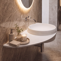 A modern wall-mounted white vessel sink in a bathroom with warm gray marble walls and floor, featuring an illuminated round mirror, chrome faucet, soap dispenser, folded towels, and a glass shower enclosure in the background.