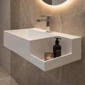 A modern floating white rectangular bathroom sink with a chrome faucet, mounted on a textured gray stone wall beneath a glowing round mirror. A small plant and folded towels sit on a wooden stool in the foreground.