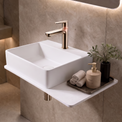A modern wall-mounted bathroom sink with a rectangular white basin and a matte brass faucet, shown installed on a beige stone wall with warm ambient lighting.