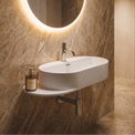 A modern wall-mounted oval bathroom sink with a chrome faucet, set against a warm beige marble wall with an illuminated round mirror and a small bottle on the shelf.