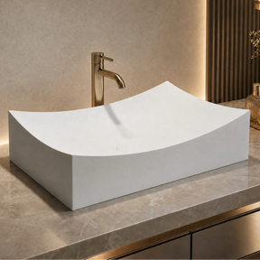 White Solid Surfaces sink on a marble countertop with a gold faucet in a bathroom setting.