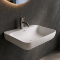 A modern wall-mounted white sink with a chrome faucet in a dark gray bathroom, featuring a large oval mirror with warm lighting and a large window overlooking a waterfall landscape.