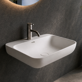 A modern wall-mounted white sink with a chrome faucet in a dark gray bathroom, featuring a large oval mirror with warm lighting and a large window overlooking a waterfall landscape.