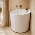 A modern freestanding oval bathtub in matte white, set in a warm beige stone bathroom with a matching faucet and small toiletry bottles on the rim. An open doorway reveals a sunlit outdoor terrace with columns, a curtain, and greenery.