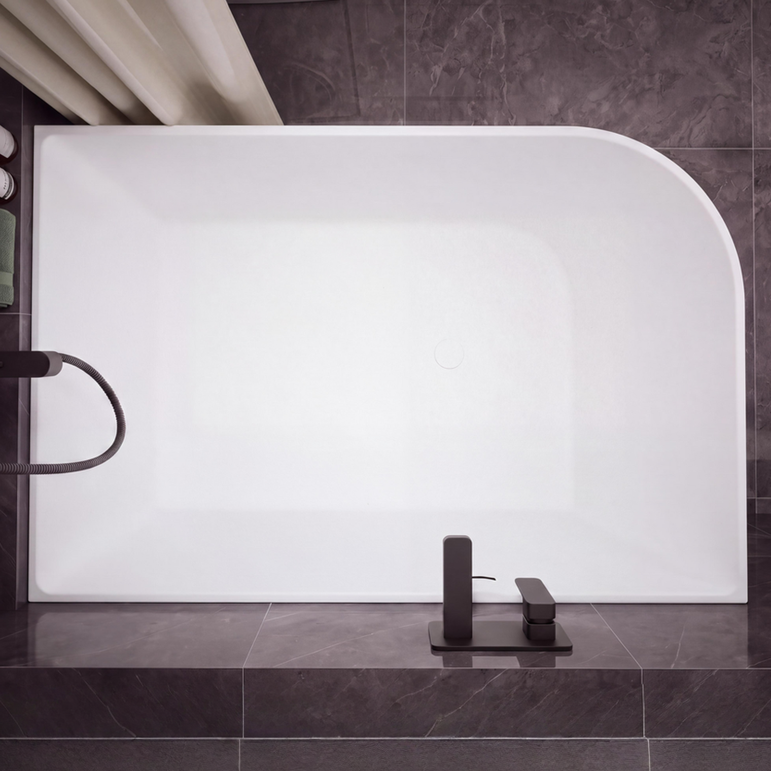 A modern freestanding rectangular bathtub with rounded corners, shown installed against dark gray tiled walls and floor. The tub is matte white, with matte black fixtures mounted on the right side and a matching black faucet centered at the bottom.