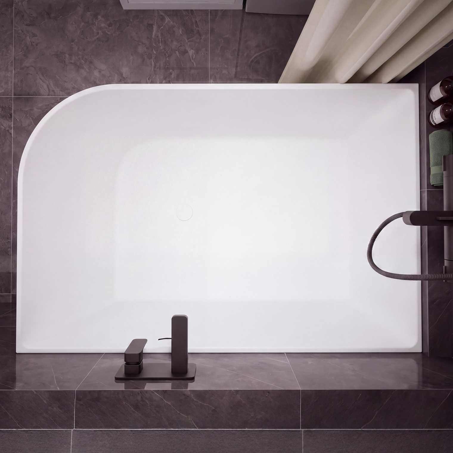 A modern freestanding rectangular bathtub with rounded corners, shown installed against dark gray tiled walls and floor. The tub is matte white, with matte black fixtures mounted on the right side and a matching black faucet centered at the bottom.