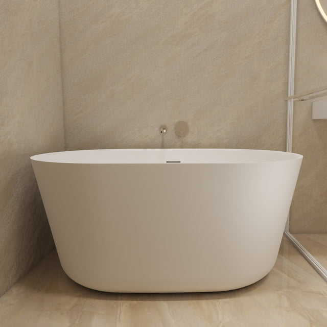 A modern freestanding oval bathtub in matte white, set in a minimalist bathroom with beige stone walls and floor, next to a glass shower screen.