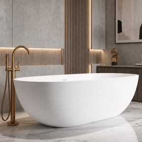 A modern freestanding oval bathtub in white with a brushed gold faucet, set in a contemporary bathroom with gray stone walls and marble flooring.