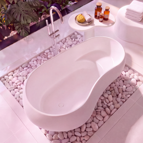 A modern freestanding oval bathtub in matt white, set on a bed of white pebbles in a bright, minimalist bathroom with tropical plants and white fixtures.