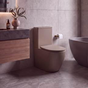 A modern bathroom interior featuring a matte taupe ceramic toilet, a floating wooden vanity with a dark stone sink, a freestanding bathtub, and large textured gray stone tiles.