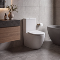 A modern bathroom interior featuring a white wall-mounted toilet next to a dark freestanding bathtub, with gray stone tile walls and floor, a floating wood-and-stone vanity, and a large window.
