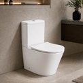 A modern white 2-piece ceramic toilet with a closed lid, shown installed in a minimalist bathroom with beige walls, a floating dark wood vanity, and a gray stone countertop.