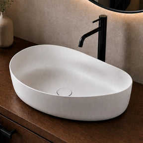 White Solid Surfaces oval sink on a wooden vanity with a black faucet, towel, and decorative items.