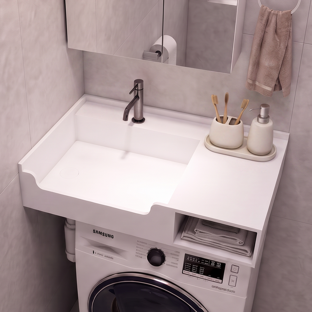 A compact bathroom vanity setup with a white sink and countertop, integrated washing machine underneath, shown in a tiled bathroom. A mirror, faucet, soap dispenser, and toilet paper holder are visible.