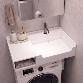 A compact bathroom vanity setup with a white sink and countertop, integrated washing machine underneath, shown in a tiled bathroom. A mirror, faucet, soap dispenser, and toilet paper holder are visible.
