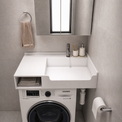 A white wall-mounted laundry sink and washer combo unit in a modern gray tiled bathroom. The white countertop features a rectangular sink basin, a side storage shelf with folded towels, a chrome faucet, and a built-in Samsung washing machine underneath.
