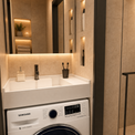 A modern bathroom vanity setup featuring a white Samsung washing machine integrated beneath a white sink with a chrome faucet, set against warm beige stone walls with built-in shelving and a glass shower enclosure.
