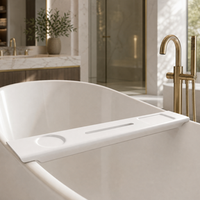 A white freestanding bathtub with a white caddy across the rim, shown in a luxurious modern bathroom with gold fixtures and marble surfaces.