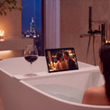 A woman sits in a white freestanding bathtub in a modern bathroom, watching a movie on a tablet placed on the tub edge. A glass of red wine sits beside the tablet, and a wall-mounted chrome faucet is visible in the background.