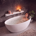 A woman relaxes in a white freestanding bathtub filled with warm water in a modern bathroom with dark gray marble walls and floor. A floating shelf holds candles and a jar of bath salts, and a potted plant sits to the side.