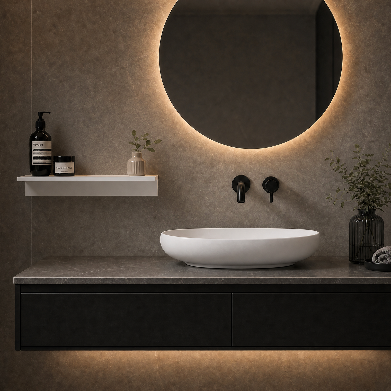 Modern bathroom vanity setup with a white oval vessel sink on a dark floating countertop, a round backlit mirror, wall-mounted black faucets, a floating shelf with toiletries, and warm ambient lighting.